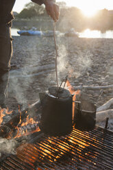 Ein Mann prüft eine Kanne Kaffee auf einem Gestell über einem Lagerfeuer bei einem Campingausflug am Fluss - MINF09923