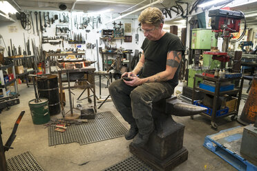 Male Blacksmith checking his phone for messages while sitting on an anvil in his studio. - MINF09984