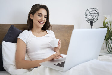 Frau mit Kaffee im Bett und Laptop, lachend - BSZF00820