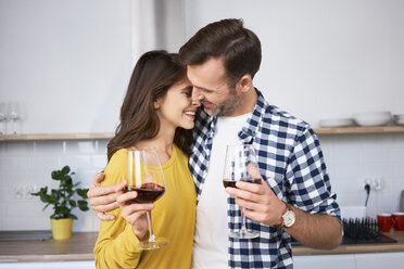 Happy couple standing in kitchen, with arms around, drinking red wine, kissing - BSZF00851