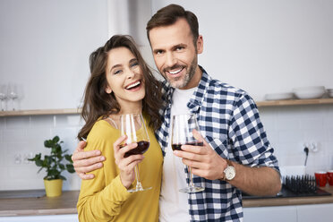 Happy couple standing in kitchen, with arms around, drinking red wine - BSZF00852