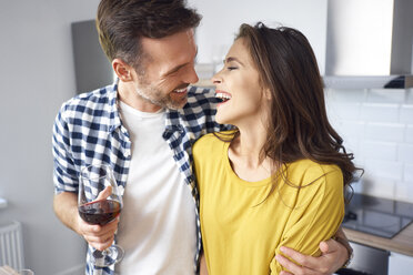 Happy couple standing in kitchen, with arms around, drinking red wine - BSZF00862