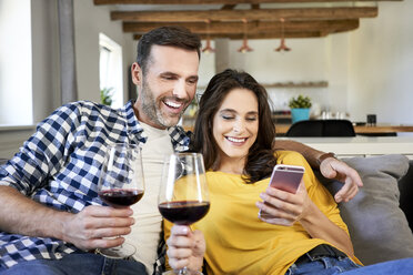 Paar sitzt im Wohnzimmer, trinkt Rotwein und benutzt ein Smartphone - BSZF00865