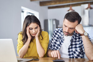 Worried couple sitting at dining table, using laptop, checking finances - BSZF00875