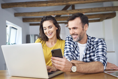 Paar sitzt am Esstisch, benutzt einen Laptop und überprüft sein Smartphone, lizenzfreies Stockfoto