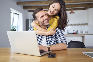 Glückliches Paar, das am Esstisch sitzt, sich umarmt und einen Laptop benutzt - BSZF00880