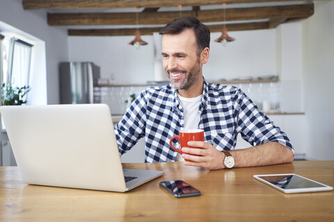 Lächelnder Mann, der zu Hause aus der Ferne an seinem Laptop arbeitet und Kaffee trinkt, lizenzfreies Stockfoto