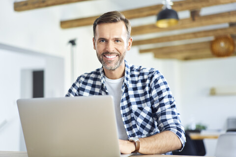 Porträt eines Mannes, der aus der Ferne an einem Laptop arbeitet und in die Kamera schaut, lizenzfreies Stockfoto