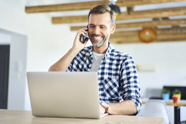 Lächelnder Mann, der am Telefon spricht, während er von zu Hause aus am Laptop arbeitet - BSZF00887