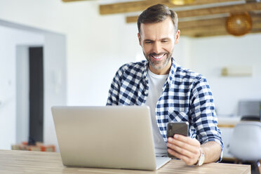 Cheerful man using phone while working on laptop at home - BSZF00889