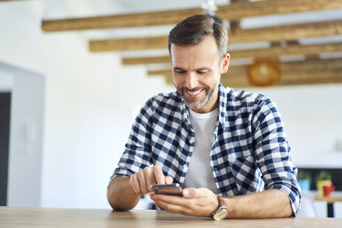 Lächelnder Mann, der zu Hause sitzt und sein Smartphone benutzt - BSZF00890