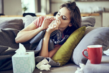 Portrait of young woman lying sick with the flu on sofa and blowing nose - BSZF00894