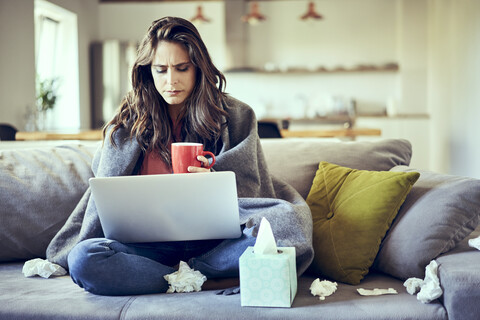 Sick woman sitting on sofa covered in blanket with cup of tea and laptop stock photo