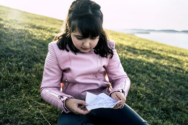 Portrait of girl wearing pink leather jacket sitting on a meadow looking at labelled paper - MGOF03937