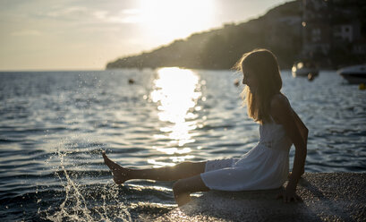 Kroatien, Lokva Rogoznica, Mädchen sitzt bei Sonnenuntergang am Wasser und spritzt mit Wasser - BFRF01959