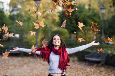 Porträt einer lachenden jungen Frau mit rotem Schal, die Herbstblätter in die Luft wirft - JSMF00732