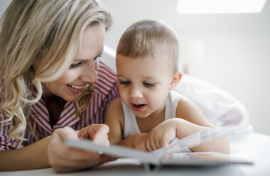 Smiling mother and toddler son lying in bed at home reading a book - HAPF02814