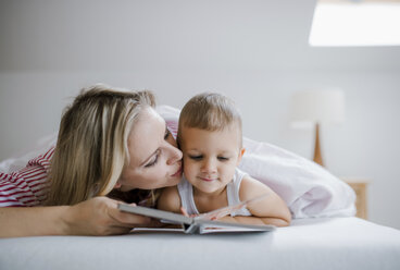 Mother kissing toddler son lying in bed at home reading a book - HAPF02815