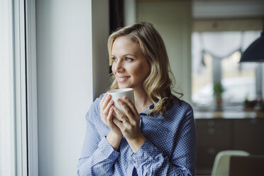 Lächelnde Frau mit einer Tasse Kaffee am Fenster zu Hause - HAPF02844