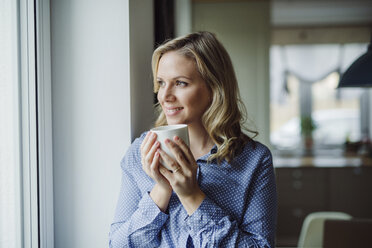 Smiling woman holding a cup of coffee at the window at home - HAPF02844