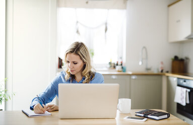 Frau mit Laptop, die zu Hause arbeitet und sich Notizen macht - HAPF02851