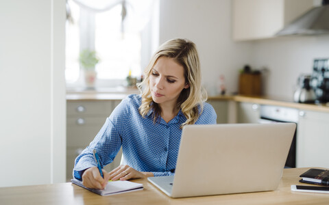 Frau mit Laptop, die zu Hause arbeitet und sich Notizen macht, lizenzfreies Stockfoto
