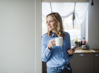 Smiling woman holding a cup of coffee and smartphone at home - HAPF02859