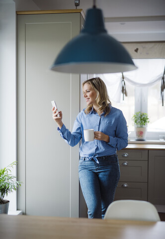 Smiling woman holding a cup of coffee and smartphone at home stock photo