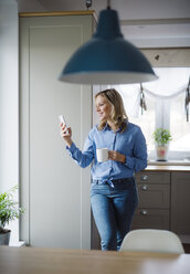 Smiling woman holding a cup of coffee and smartphone at home - HAPF02860