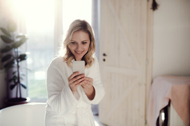 Smiling woman wearing bathrobe in bathroom at home holding cell phone - HAPF02871