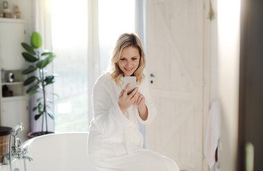 Smiling woman wearing bathrobe in bathroom at home holding cell phone - HAPF02872