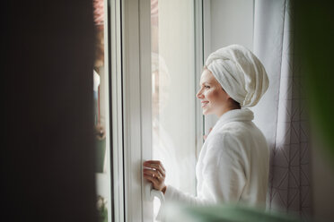 Lächelnde Frau im Bademantel mit Handtuch um den Kopf schaut zu Hause aus dem Fenster - HAPF02874