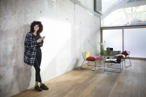 Smiling woman with cell phone leaning against concrete wall in a loft stock photo