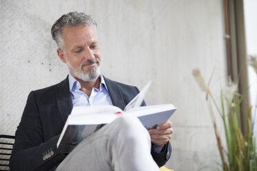 Smiling businessman sitting at concrete wall reading book - FKF03163