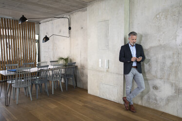Businessman using cell phone leaning against concrete wall in a loft - FKF03167