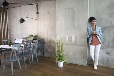 Lächelnde Geschäftsfrau hält Kaffeebecher an Betonwand in einem Loft - FKF03212