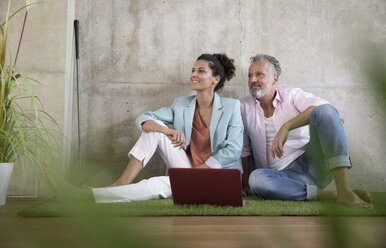 Lässiger Geschäftsmann und Geschäftsfrau sitzen auf Kunstrasen in einem Loft und teilen sich einen Laptop - FKF03222