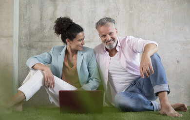 Casual businessman and businesswoman sitting on artificial turf in a loft sharing laptop - FKF03224