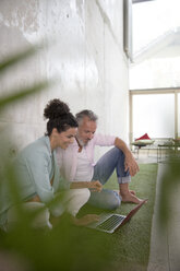 Casual businessman and businesswoman sitting on artificial turf in a loft sharing laptop - FKF03227