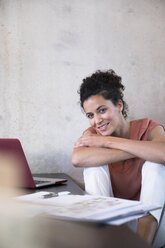 Portrait of smiling businesswoman sitting at table with documents and laptop - FKF03239