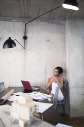 Portrait of smiling businesswoman sitting at table in a loft with documents and laptop - FKF03243