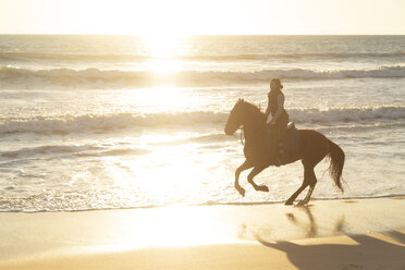 Spanien, Tarifa, Frau reitet Pferd am Strand bei Sonnenuntergang - KBF00387