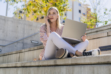 Porträt einer blonden jungen Frau, die mit einem Laptop auf einer Treppe im Freien sitzt und telefoniert - GIOF05457