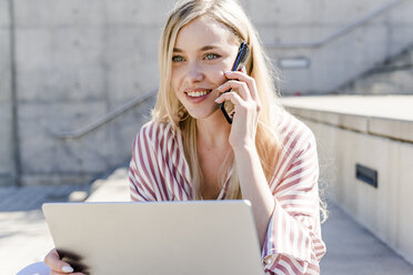Porträt einer lächelnden blonden jungen Frau mit Laptop, die im Freien telefoniert - GIOF05458