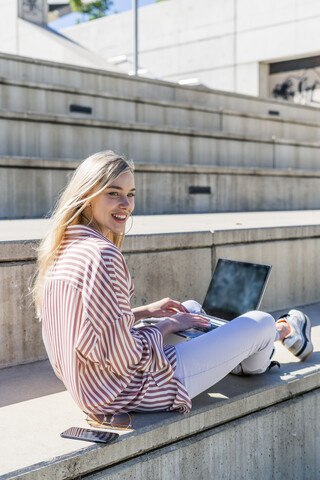Porträt einer lächelnden jungen Frau mit Laptop, die auf einer Treppe im Freien sitzt und etwas beobachtet, lizenzfreies Stockfoto