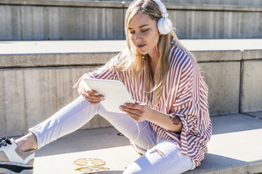 Portrait of blond young woman sitting on stairs outdoors using digital tablet and headphones - GIOF05460