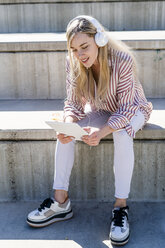 Porträt einer blonden jungen Frau, die auf einer Treppe im Freien sitzt und ein digitales Tablet und Kopfhörer benutzt - GIOF05461