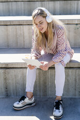 Portrait of blond young woman sitting on stairs outdoors using digital tablet and headphones - GIOF05461