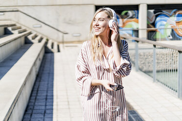 Spain, Barcelona, portrait of smiling blond woman using smartphone and headphones outdoors - GIOF05463