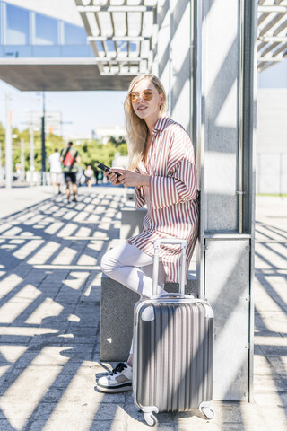 Spanien, Barcelona, junge Frau mit Trolley-Tasche und Mobiltelefon wartet am Bahnhof, lizenzfreies Stockfoto